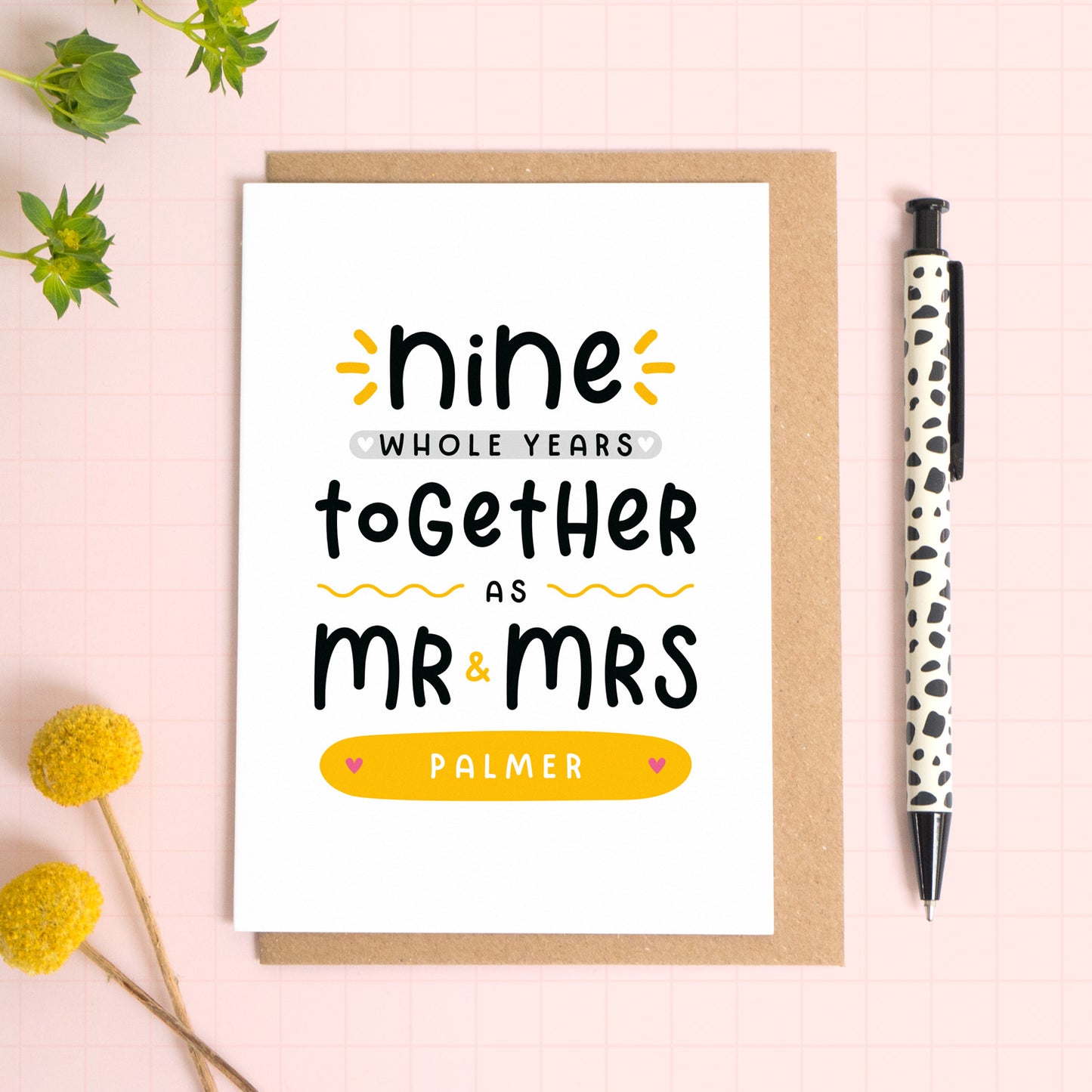 A personalised Mr & Mrs anniversary card reading ‘nine whole years together as Mr & Mrs [surname]’ which has been photographed on a Kraft brown envelope upon a pink background surrounded by florals and a pen for scale.