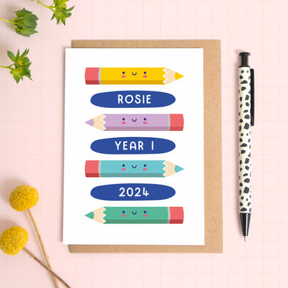 A personalised good luck going to school card featuring a lots of colourful pencils, the recipients name, school year and year. The card has been photographed on it's kraft brown envelope on a pink surface with foliage and a pen for scale. 