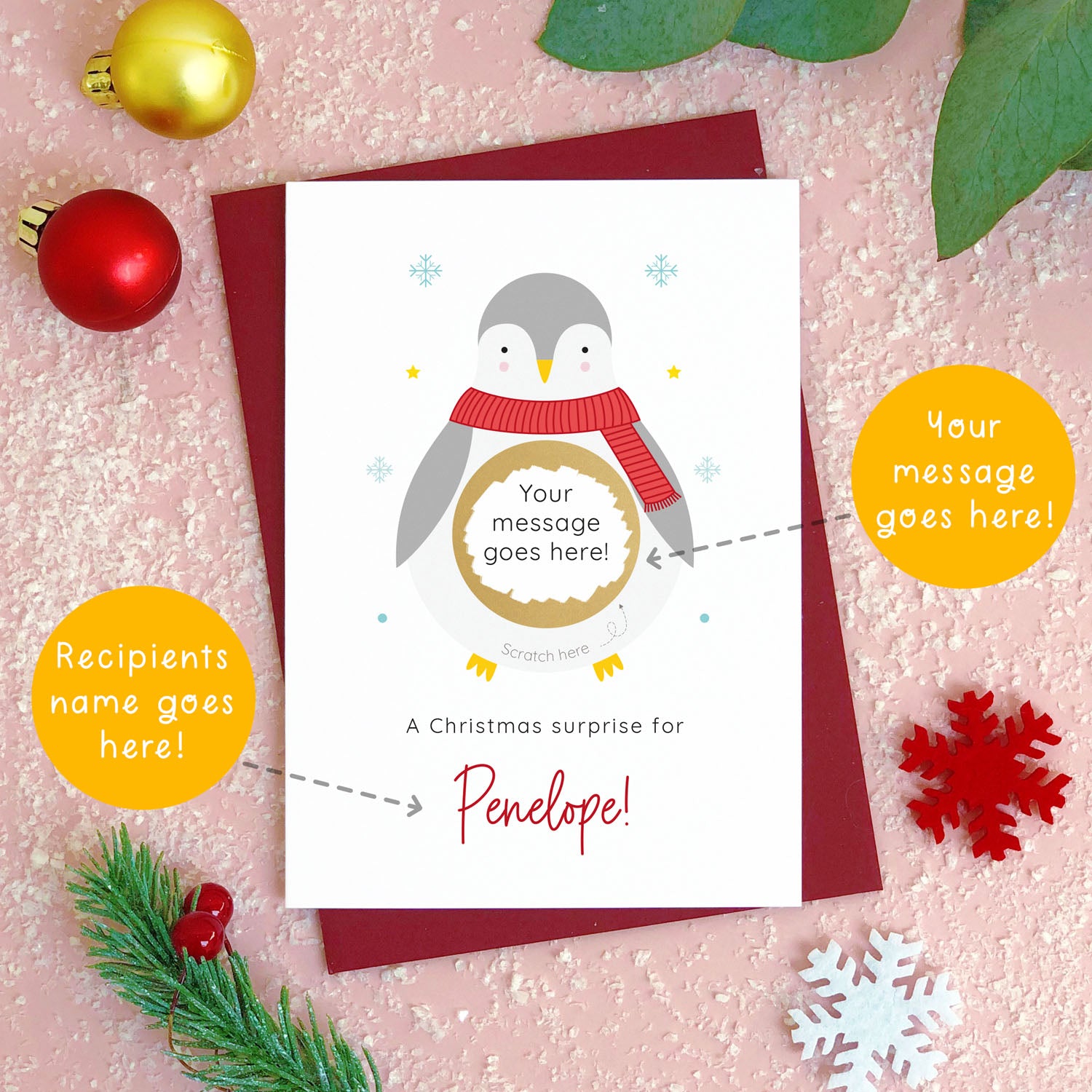 A personalised penguin Christmas scratch card photographed flat lying on a red wine coloured envelope. The orange circles demonstrate which areas of the card can be personalised. Here they point to the message and name for the front of the card.