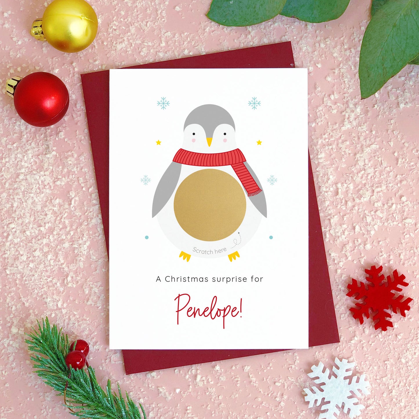 A personalised penguin Christmas scratch card photographed flat lying on a red wine coloured envelope on a pink surface surrounded by fake snow, baubles and foliage. The gold panel has not yet been scratched off to reveal the custom message.