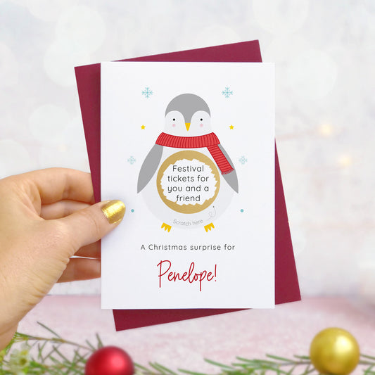A personalised penguin Christmas scratch card photographed being held by a hand to the left and foliage and baubles in the bottom of the frame. The card is against its ‘red wine’ colour envelope and the scratch off panel has been scratched off to reveal a trip to lapland!
