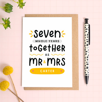 A personalised Mr & Mrs anniversary card reading ‘seven whole years together as Mr & Mrs [surname]’ which has been photographed on a Kraft brown envelope upon a pink background surrounded by florals and a pen for scale.