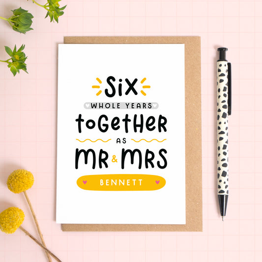 A personalised Mr & Mrs anniversary card reading ‘six whole years together as Mr & Mrs [surname]’ which has been photographed on a Kraft brown envelope upon a pink background surrounded by florals and a pen for scale.