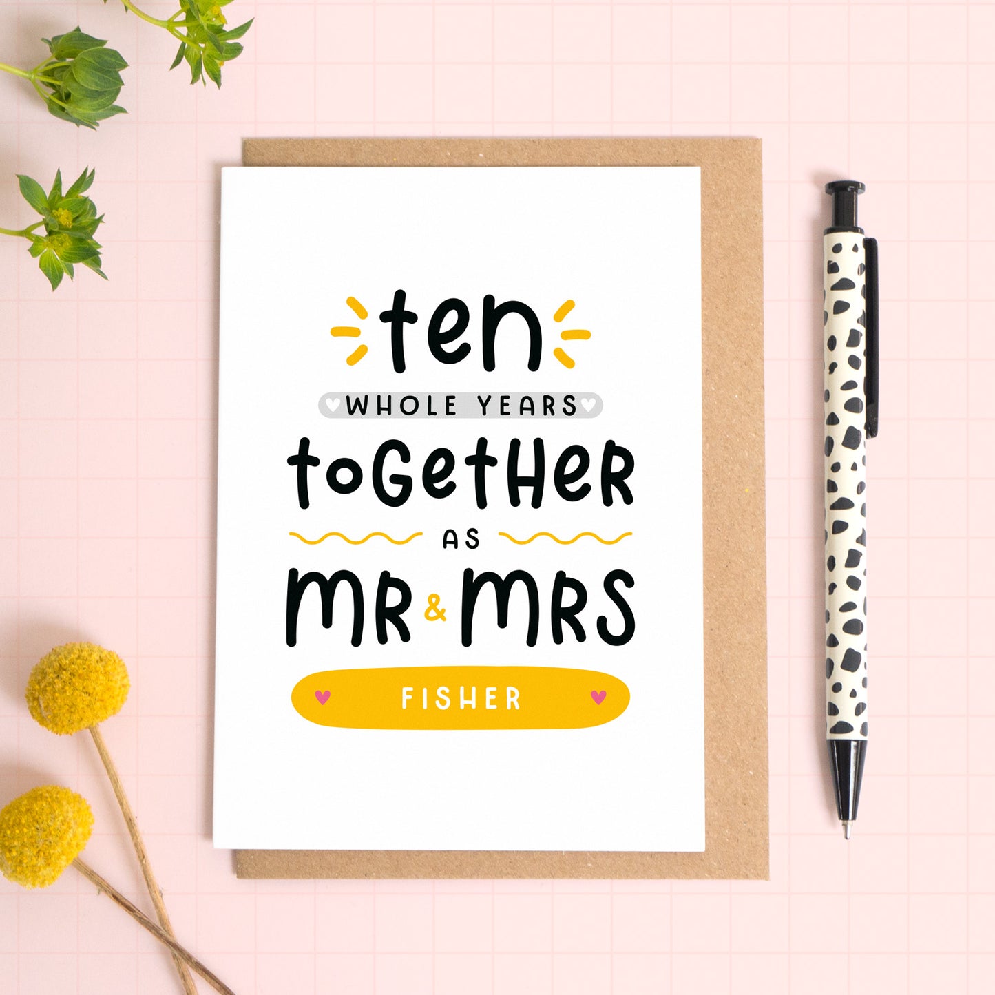 A personalised Mr & Mrs anniversary card reading ‘ten whole years together as Mr & Mrs [surname]’ which has been photographed on a Kraft brown envelope upon a pink background surrounded by florals and a pen for scale.