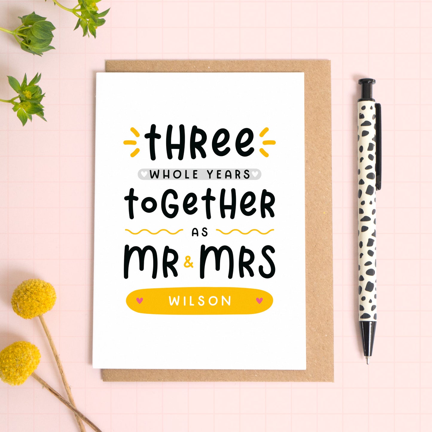 A personalised Mr & Mrs anniversary card reading ‘three whole years together as Mr & Mrs [surname]’ which has been photographed on a Kraft brown envelope upon a pink background surrounded by florals and a pen for scale.