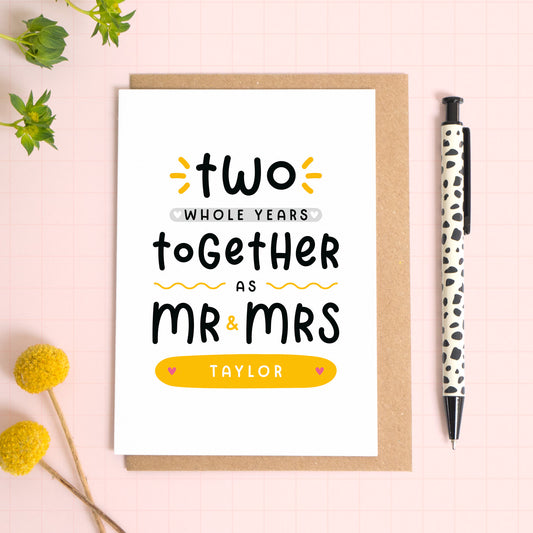 A personalised Mr & Mrs anniversary card reading ‘two whole years together as Mr & Mrs [surname]’ which has been photographed on a Kraft brown envelope upon a pink background surrounded by florals and a pen for scale.