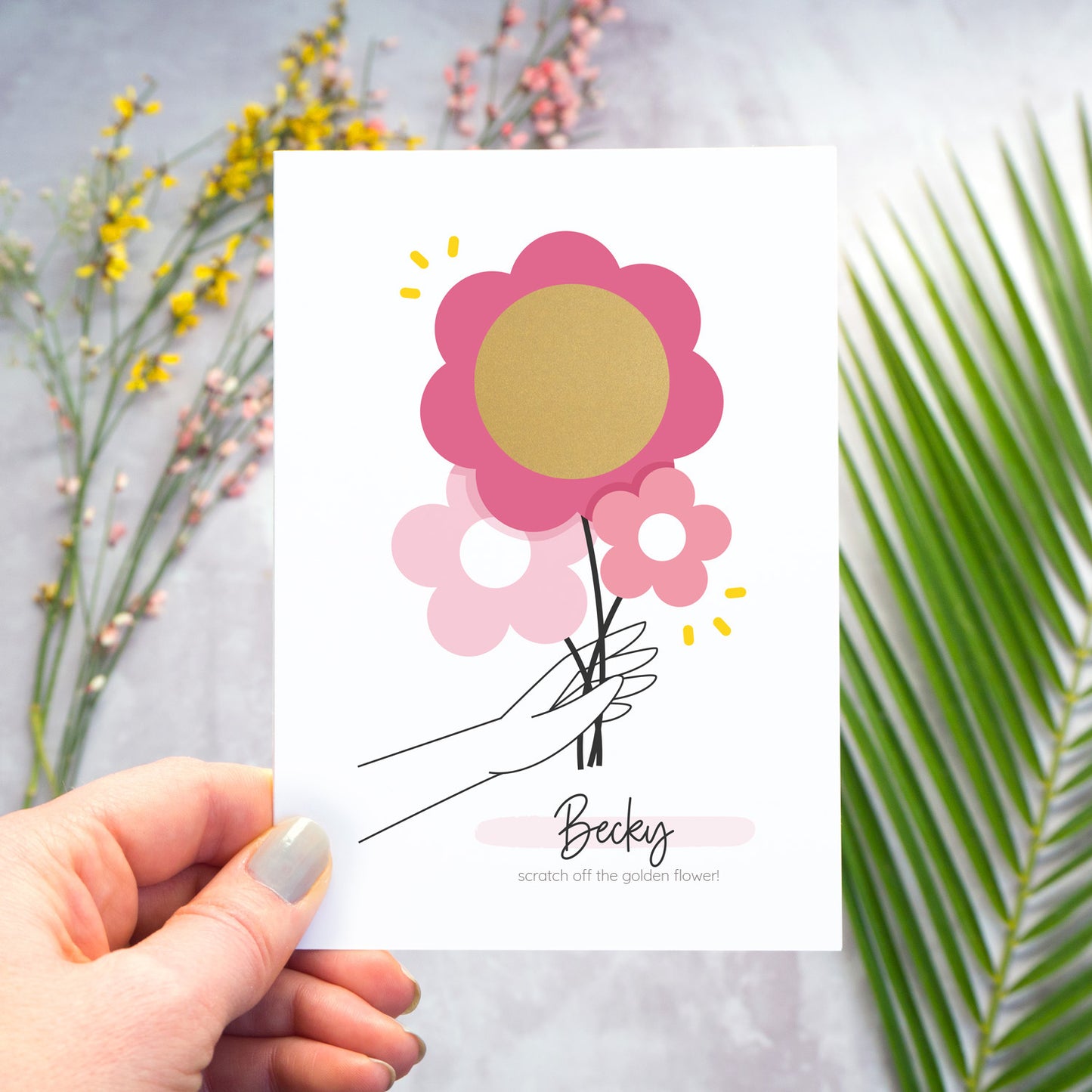 This image shows how the personalised question scratch card looks before the recipient scratches away the flowers inner circle. In this image the card is being held over a grey surface which has a variety of colour flowers and foliage in the background. The heart has not yet been scratched away.