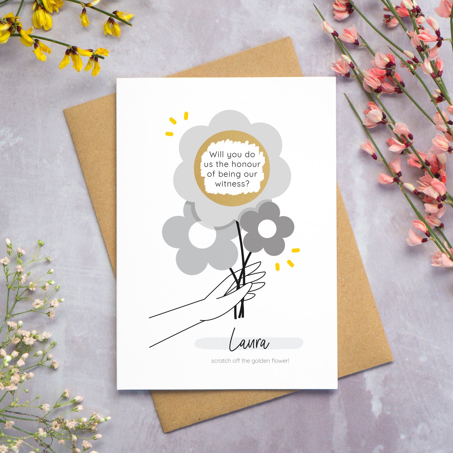 The personalised question scratch card has been photographed lying on top of a Kraft brown envelope which is on top of a grey surface surrounded by colourful flowers. The gold circle scratch panel has been scratched away to reveal the surprise question. This is the grey colour palette.
