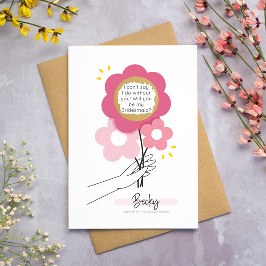 The personalised question scratch card has been photographed lying on top of a Kraft brown envelope which is on top of a grey surface surrounded by colourful flowers. The gold circle scratch panel has been scratched away to reveal the surprise question. This is the pink colour palette.