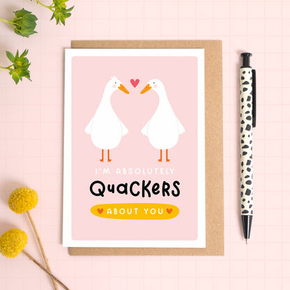 A duck pun anniversary card featuring two cute ducks and the text ‘I’m absolutely Quackers about you’, which has been photographed on a Kraft brown envelope upon a pink background surrounded by florals and a pen for scale.