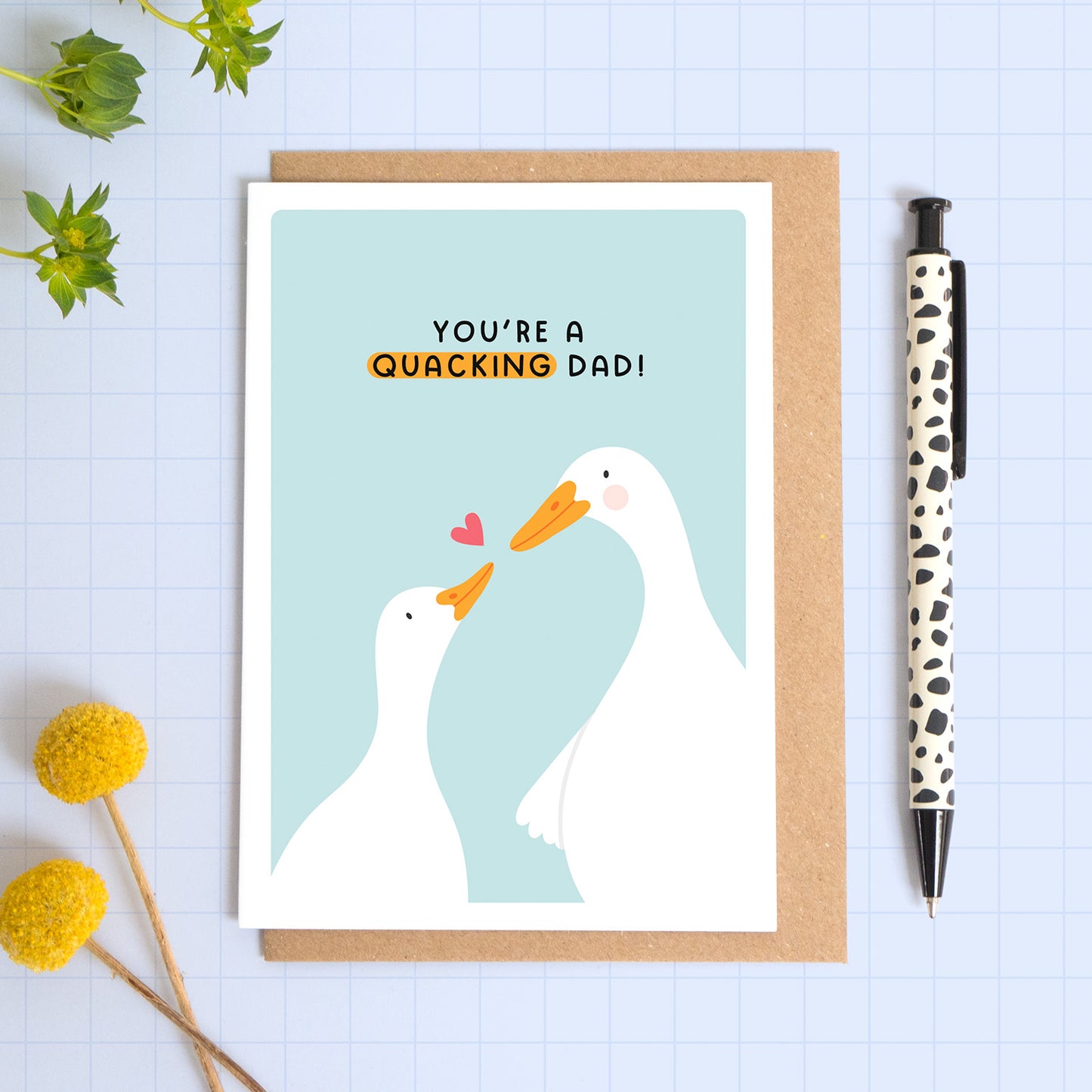 A blue card with a white border featuring two cute ducks and the phrase 'you're a quacking dad!’. The card is laid on a kraft brown envelope on top of a blue background next to a pen and flowers.