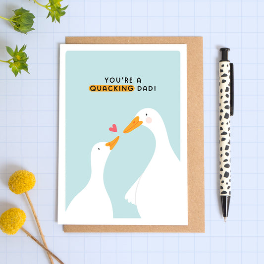 A blue card with a white border featuring two cute ducks and the phrase 'you're a quacking dad!’. The card is laid on a kraft brown envelope on top of a blue background next to a pen and flowers.