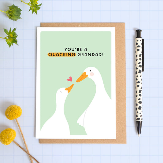 A green card with a white border featuring two cute ducks and the phrase 'you're a quacking grandad!’. The card is laid on a kraft brown envelope on top of a blue background next to a pen and flowers.