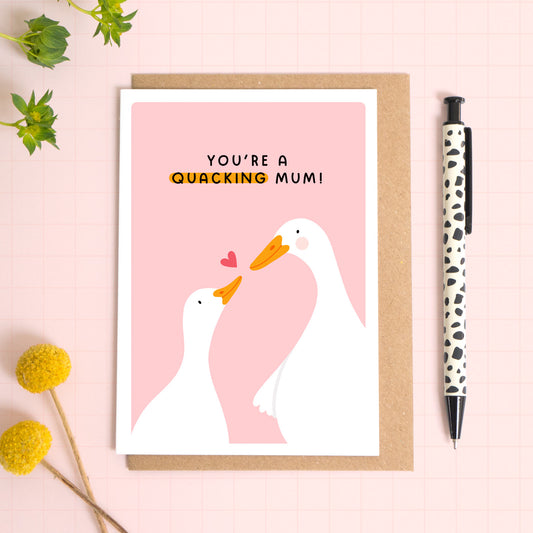 A pink card with a white border featuring two cute ducks and the phrase 'you're a quacking mum!'. The card is laid on a kraft brown envelope on top of a pink background next to a pen and flowers.