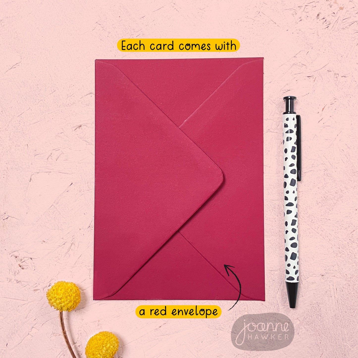 A red envelope that comes with each of the greetings card. It has been photographed on a pink background next to yellow flowers and a pen for scale.