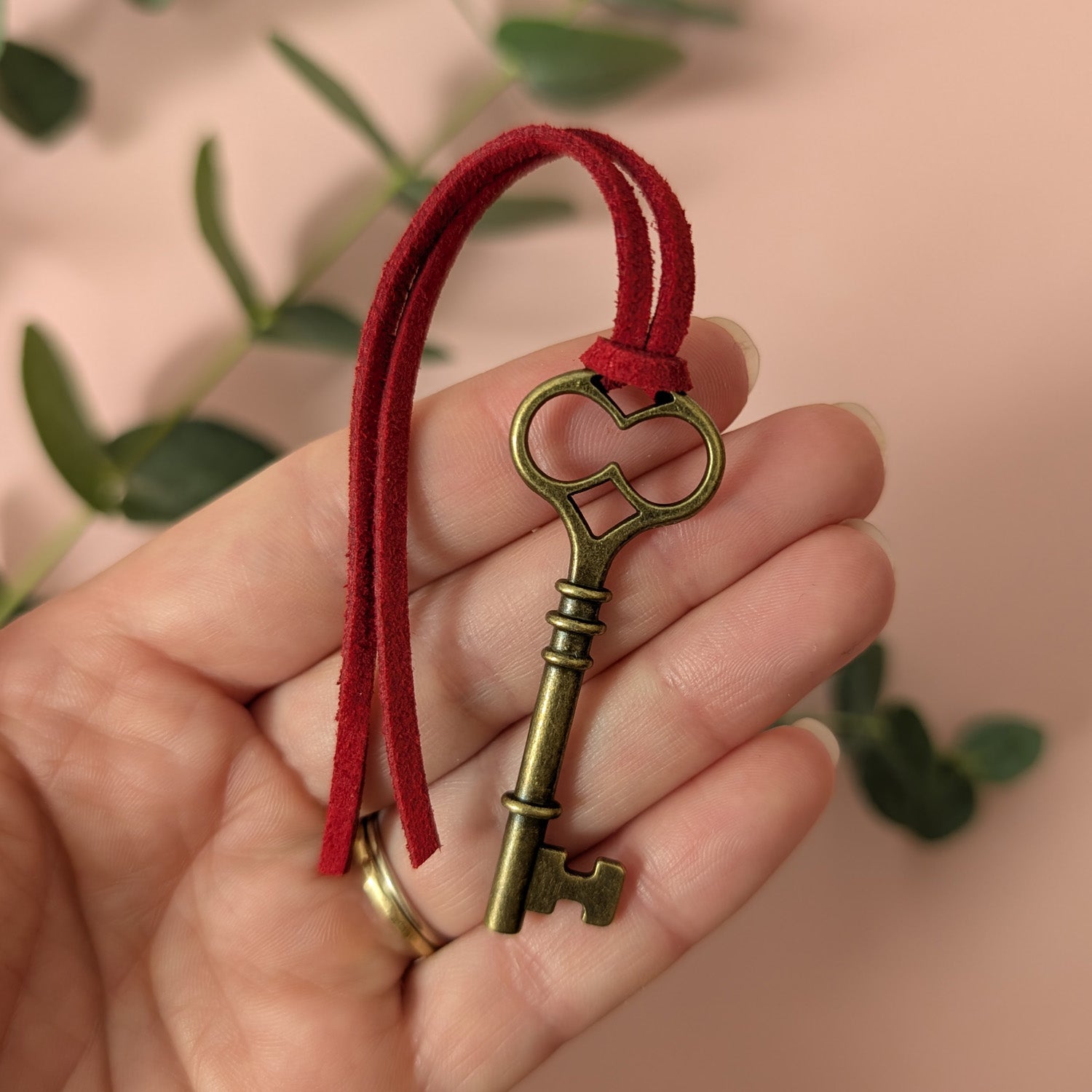 An antique style themed key with red string held in a hand against a blurred green leafy background.
