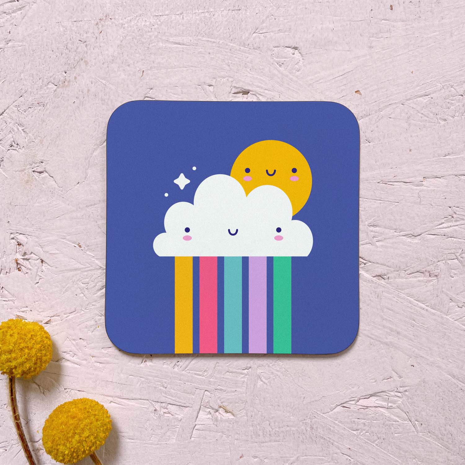 A single rainbow cloud themed coaster featuring a happy sun, a smiling cloud and a rainbow. It has been photographed on a pink background with two yellow flowers.