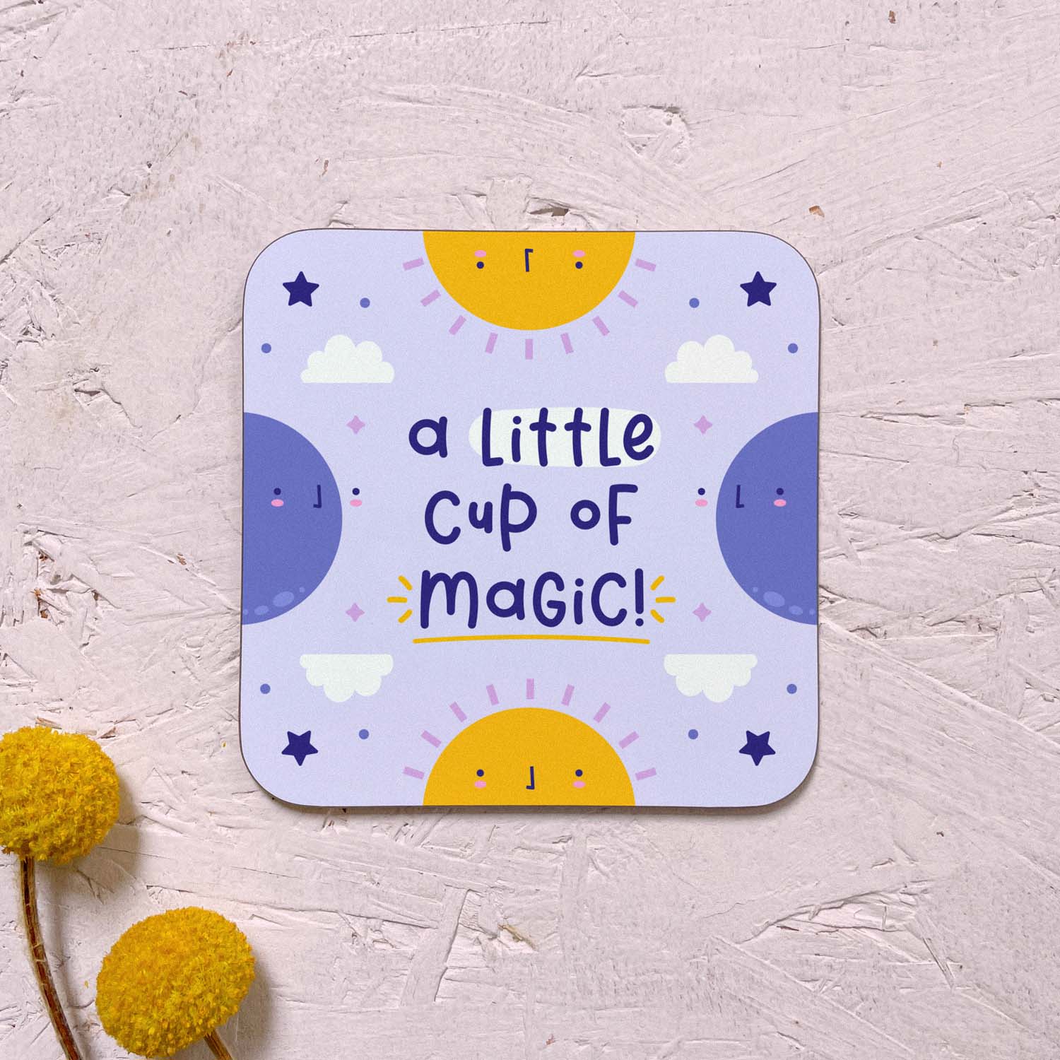 A single celestial themed coaster featuring yellow suns, blue moons, stars and clouds and the text 'a little cup of magic!'. It has been photographed on a pink background with two yellow flowers.