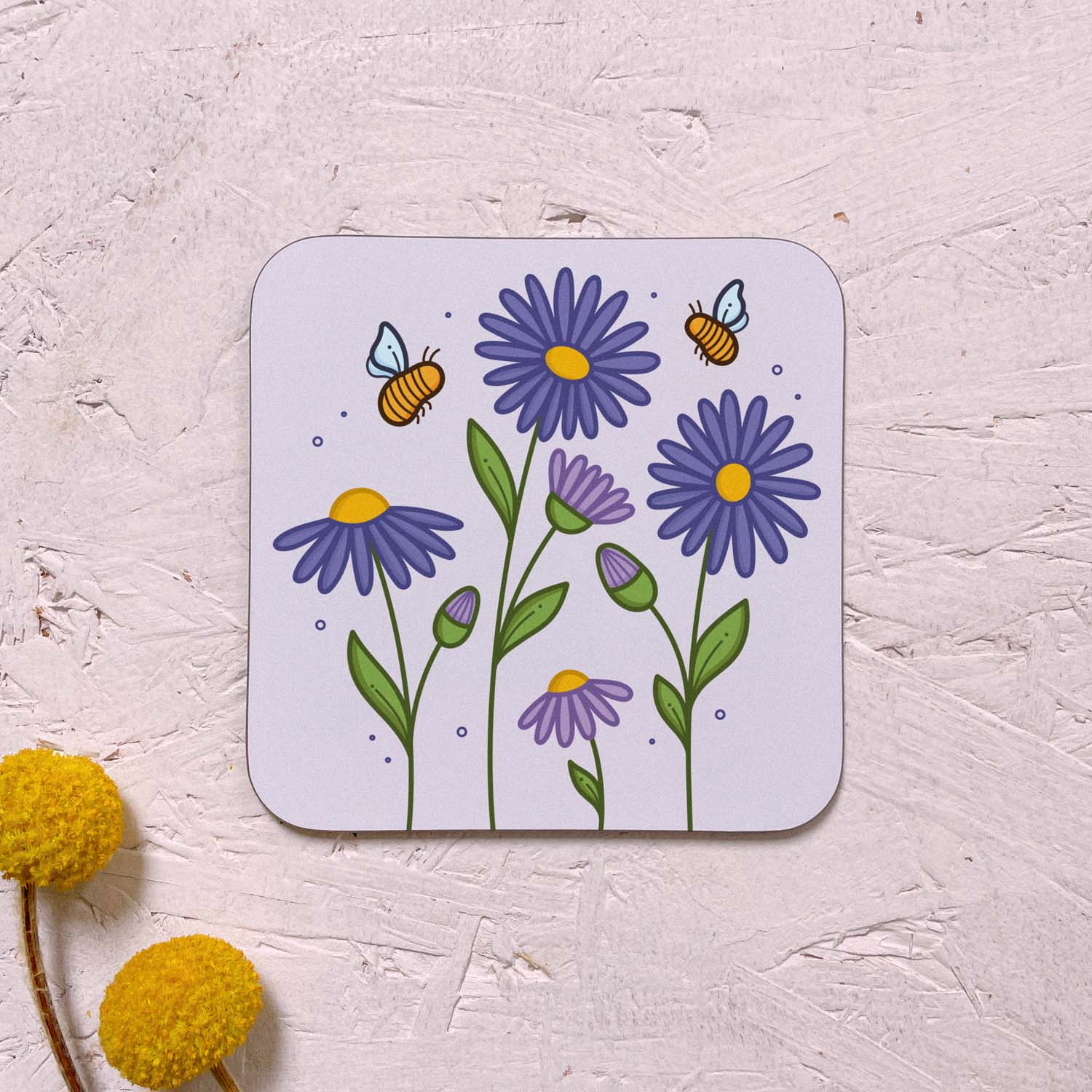 A single floral themed coaster featuring purple aster flowers, and yellow bumble bees on a lilac grey background. It has been photographed on a pink background with two yellow flowers.