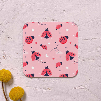 A single ladybug cloud themed coaster featuring a variety of ladybirds, stars and clouds. It has been photographed on a pink background with two yellow flowers.