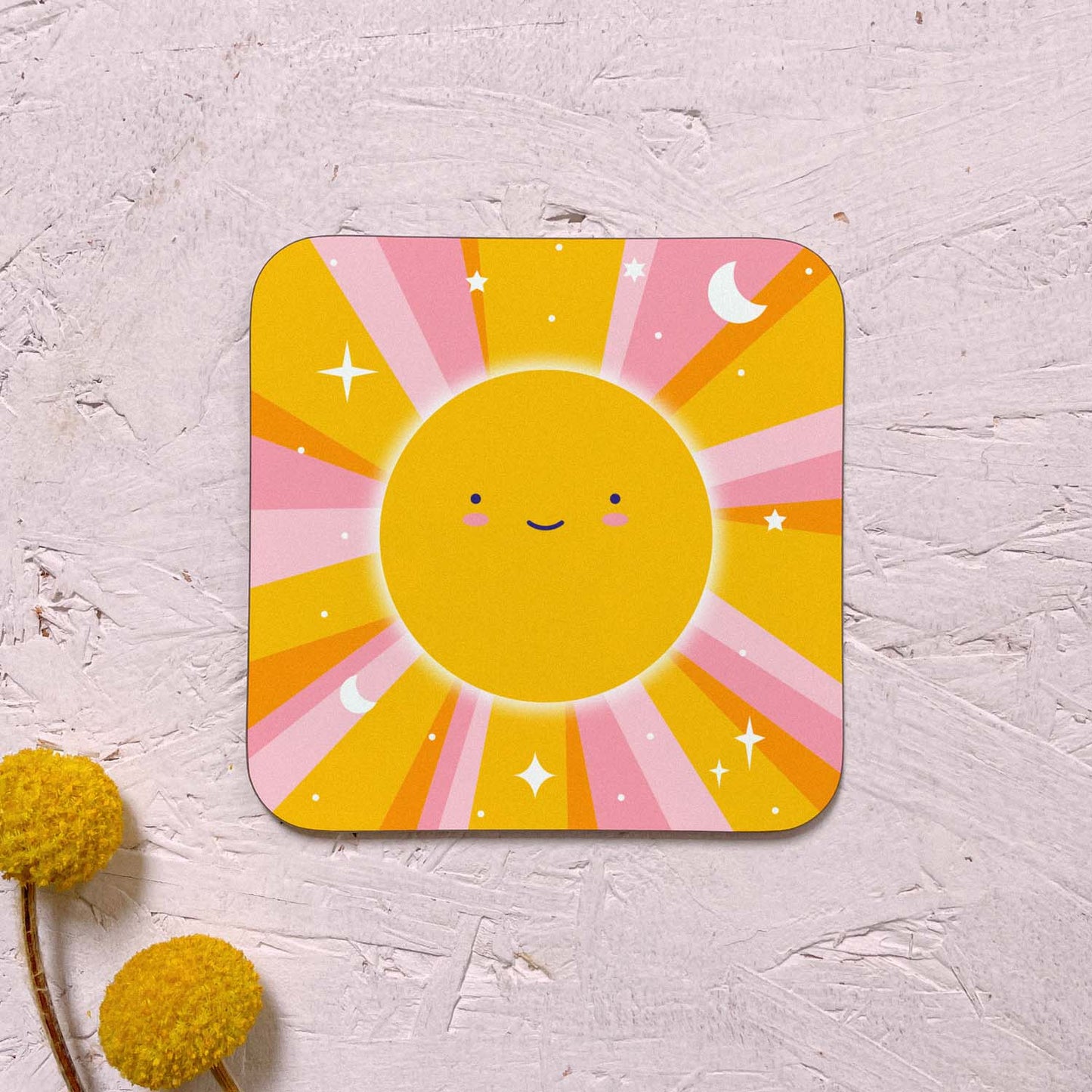 A single celestial sun themed coaster featuring a smiling happy sun with yellow and pink sunbeams littered with stars, moons and dots. It has been photographed on a pink background with two yellow flowers.