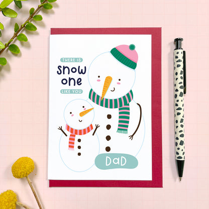 A Christmas card for a lovely dad featuring a small snowman looking up at a bigger one with the text 'there is snow one like you dad.' The card has a red envelope and it has been photographed on a pink background next to yellow flowers and a pen for scale.