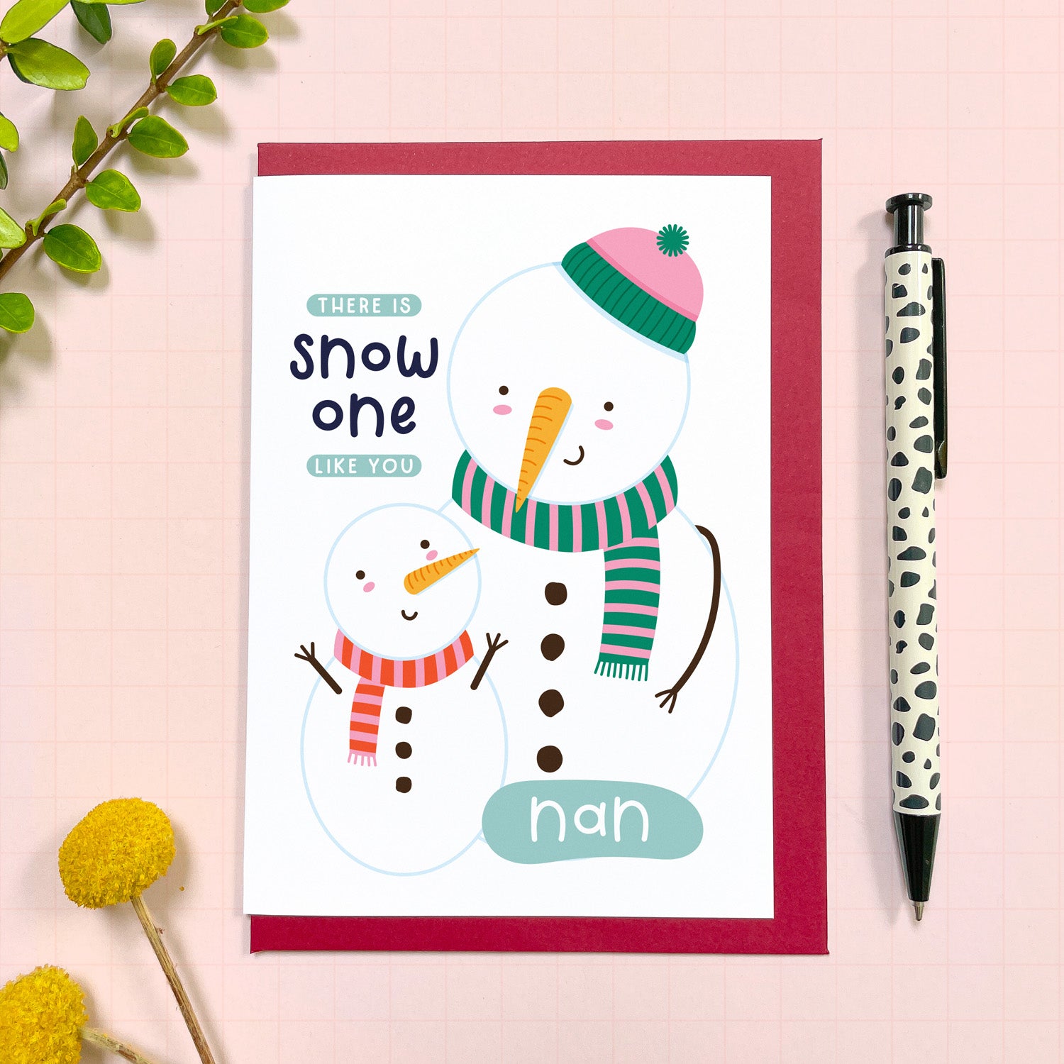 A Christmas card for a lovely nan featuring a small snowman looking up at a bigger one with the text 'there is snow one like you nan.' The card has a red envelope and it has been photographed on a pink background next to yellow flowers and a pen for scale.