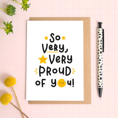 A very proud of you card featuring the text "so very, very proud of you!' and a star and sun with smiling faces. The card has been photographed lying on top of a kraft brown envelope with foliage to the side and a pen for scale.