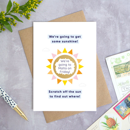 A holiday reveal scratch card featuring a shiny gold sun. The gold panel has been scratched off to reveal the holiday destination. The text reads: “We're going to get some sunshine! Scratch off the sun to find out where!”. This card has been shot on a grey background with a pen and foliage. 