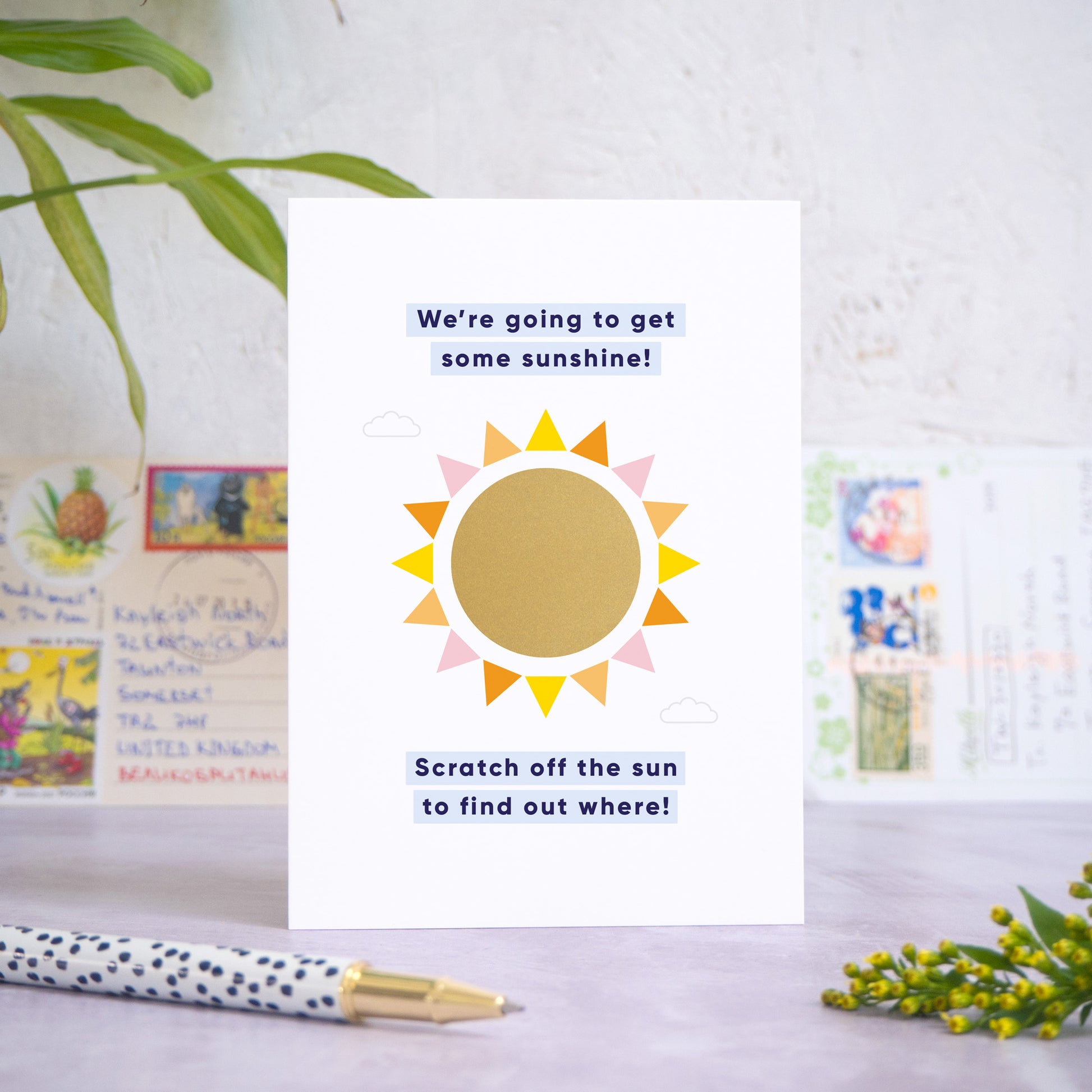 A holiday reveal scratch card featuring a shiny gold sun. The gold panel has not yet been scratched off to reveal the holiday destination. The text reads: “We're going to get some sunshine! Scratch off the sun to find out where!”. The card is standing on a grey surface with postcards in the background and a pen and foliage in the foreground.