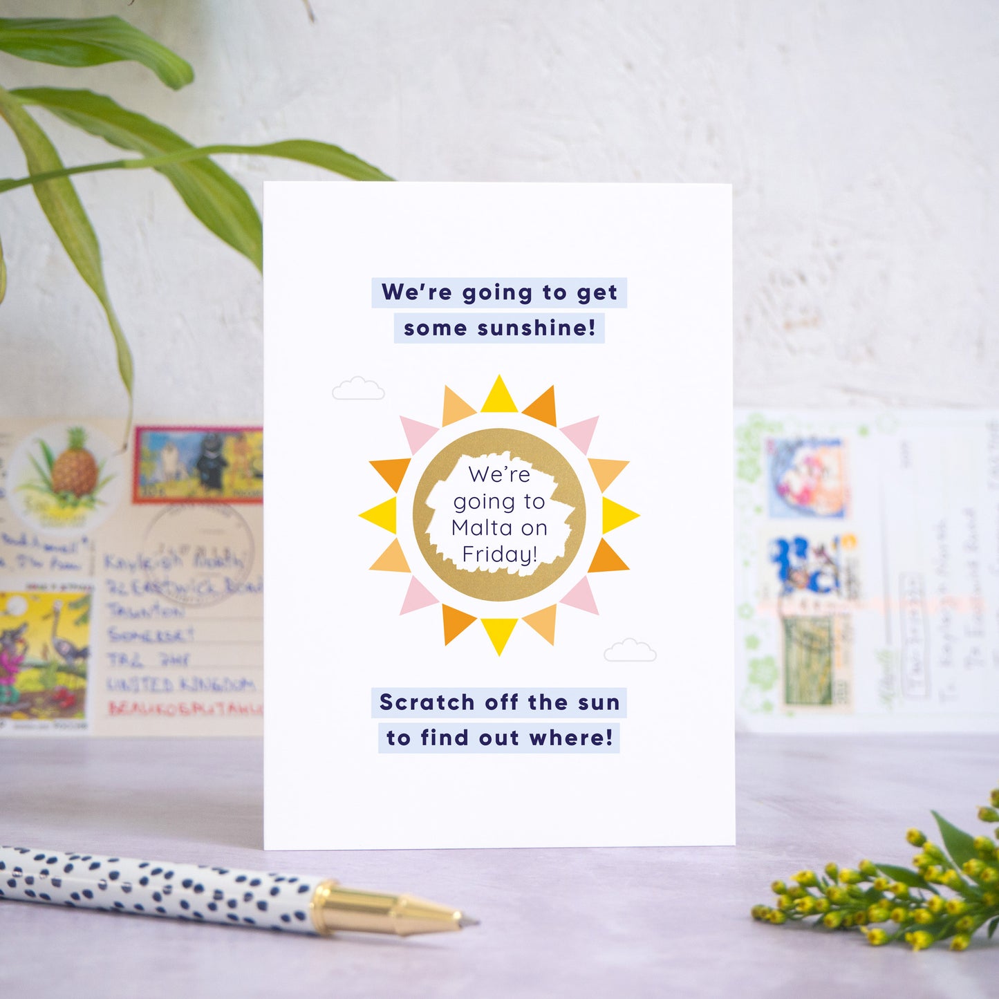 A holiday reveal scratch card featuring a shiny gold sun. The gold panel has been scratched off to reveal the holiday destination. The text reads: “We're going to get some sunshine! Scratch off the sun to find out where!”. The card is standing on a grey surface with postcards in the background and a pen and foliage in the foreground.
