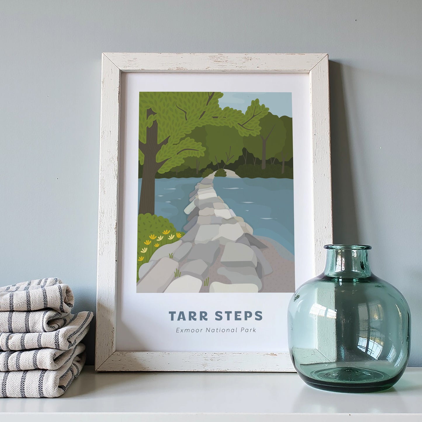 Tarr Steps, Exmoor National Park art print in a white frame on a light grey background with a pile of cloths and blue vase.
