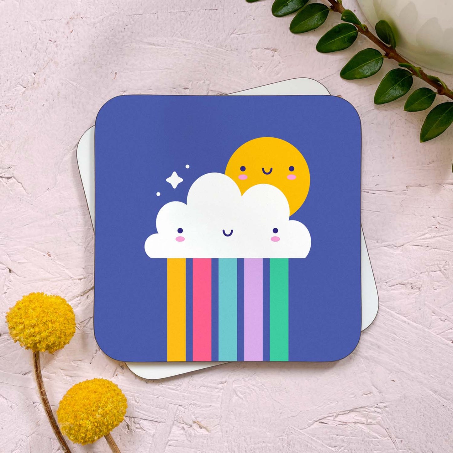 A cloud and rainbow themed coaster that has been photographed stacked on top of a blank coaster on a pink background. To the top right is a sprig of greenery and a mug, and in the bottom left are two flowers. This design features a smiling sun hiding behind a happy cloud with a colourful rainbow raining out from beneath.