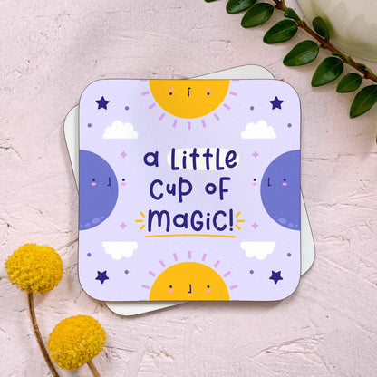 A celestial themed coaster that has been photographed stacked on top of a blank coaster on a pink background. To the top right is a sprig of greenery and a mug, and in the bottom left are two flowers. The celestial design features two suns, two moons, stars and clouds with the text 'a little cup of magic!'