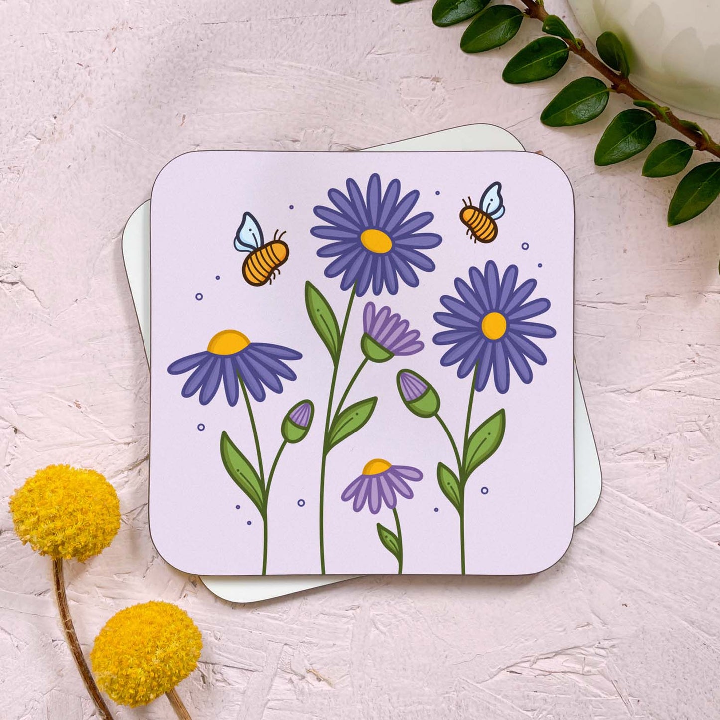 A floral themed coaster that has been photographed stacked on top of a blank coaster on a pink background. To the top right is a sprig of greenery and a mug, and in the bottom left are two flowers. The floral design features purple aster flowers and two yellow bumble bees on top of a lilac grey background.