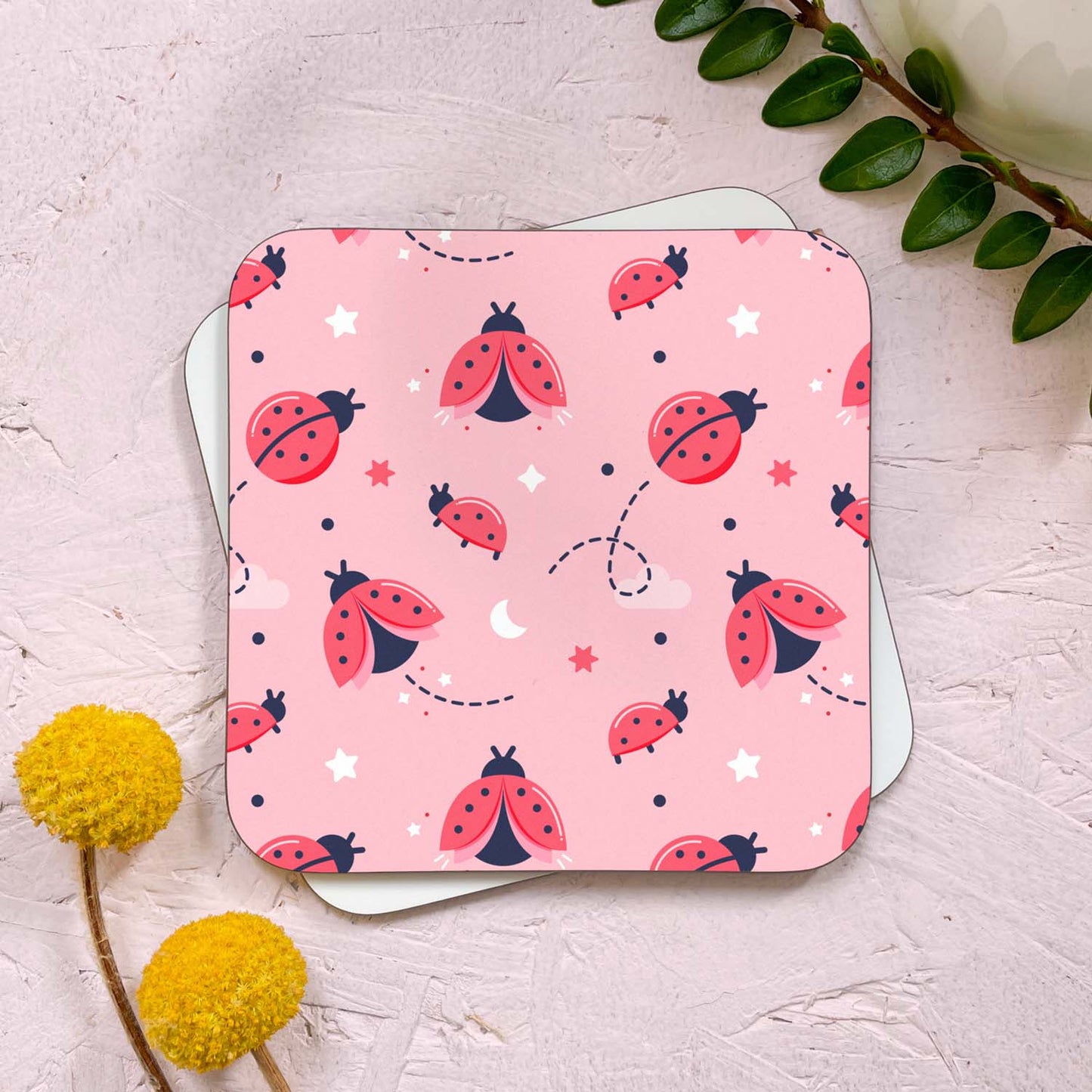 A ladybug themed coaster that has been photographed stacked on top of a blank coaster on a pink background. To the top right is a sprig of greenery and a mug, and in the bottom left are two flowers. This design features a variety of ladybirds flying on a pink background surrounded by stars and clouds.