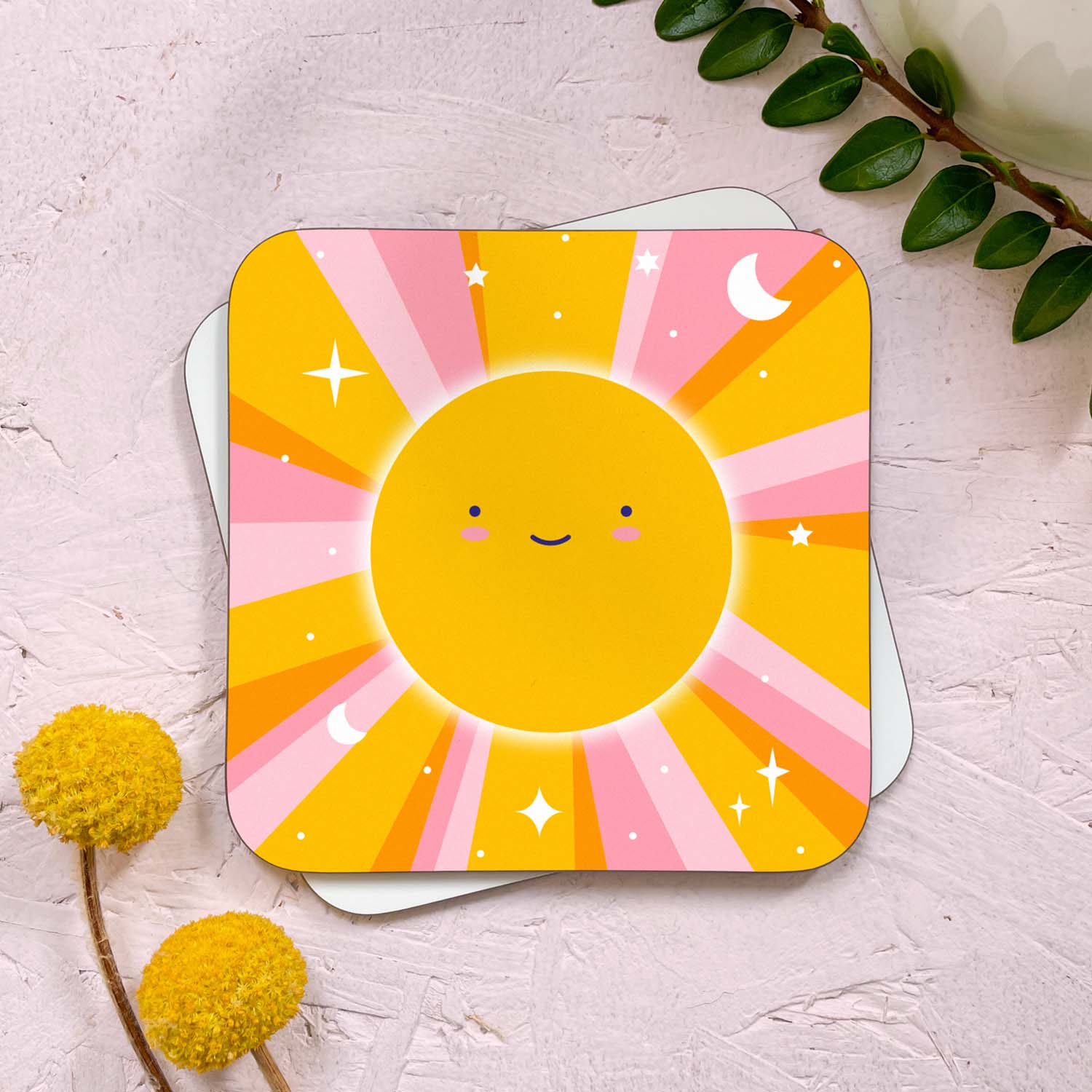 A celestial sun themed coaster that has been photographed stacked on top of a blank coaster on a pink background. To the top right is a sprig of greenery and a mug, and in the bottom left are two flowers. The celestial sun design features a smiling sun, pink and yellow sunbeams littered with stars, moons and dots.