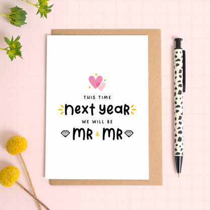 A last anniversary before marriage card with overlapping hearts and text that reads ‘This time next year we will be Mr and Mr’. This wedding count down card has been photographed on a Kraft brown envelope upon a pink background with a pen for scale.