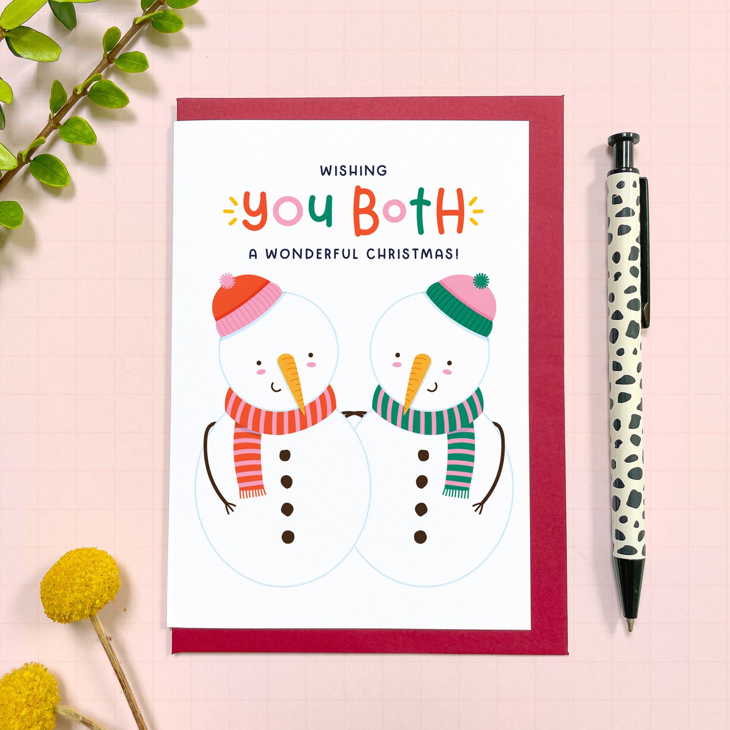 A 'for the both of you' themed Christmas card with two snowmen wearing hats and scarves. The card has a red envelope and it has been photographed on a pink background next to yellow flowers and a pen for scale.