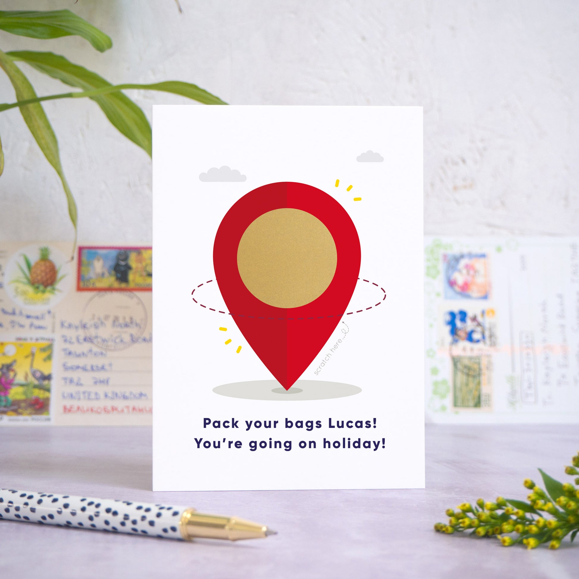 A location pin holiday reveal scratch card featuring a big red location marker, with clouds in the background. The location is in the centre of the pin and has been hidden underneath the gold panel scratch panel. Beneath the pin reads: “Pack your bags [insert recipients name]! You're going on holiday!”. The card is standing on a grey surface with postcards in the background and a pen and foliage in the foreground.