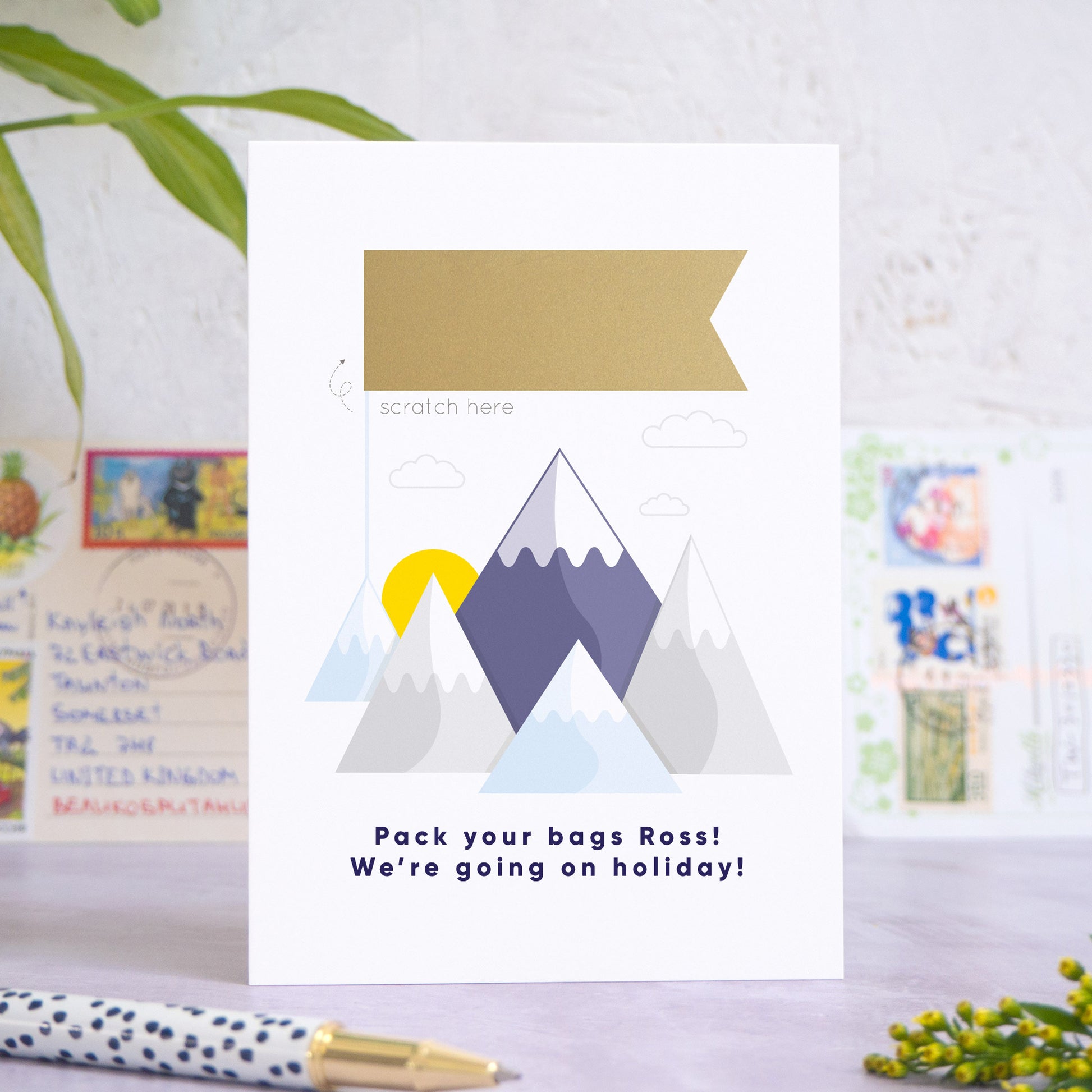 A mountain holiday reveal scratch card featuring a mountain range, with a sun peeping out behind the peaks, little clouds and a gold flag. The flag has yet to be scratched off. Beneath the mountain range reads: “Pack your bags [insert recipients name]! We're going on holiday!”. The card is standing on a grey surface with postcards in the background and a pen and foliage in the foreground.