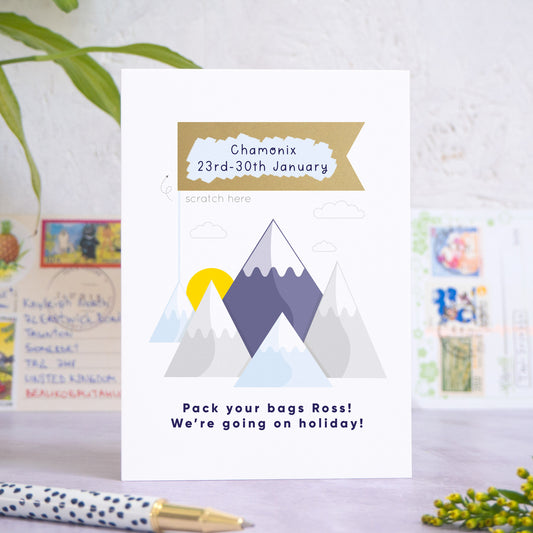 A mountain holiday reveal scratch card featuring a mountain range, with a sun peeping out behind the peaks, little clouds and a gold flag. The flag has been scratched off to reveal the details of the holiday location and the dates. Beneath the mountain range reads: “Pack your bags [insert recipients name]! We're going on holiday!”. The card is standing on a grey surface with postcards in the background and a pen and foliage in the foreground.