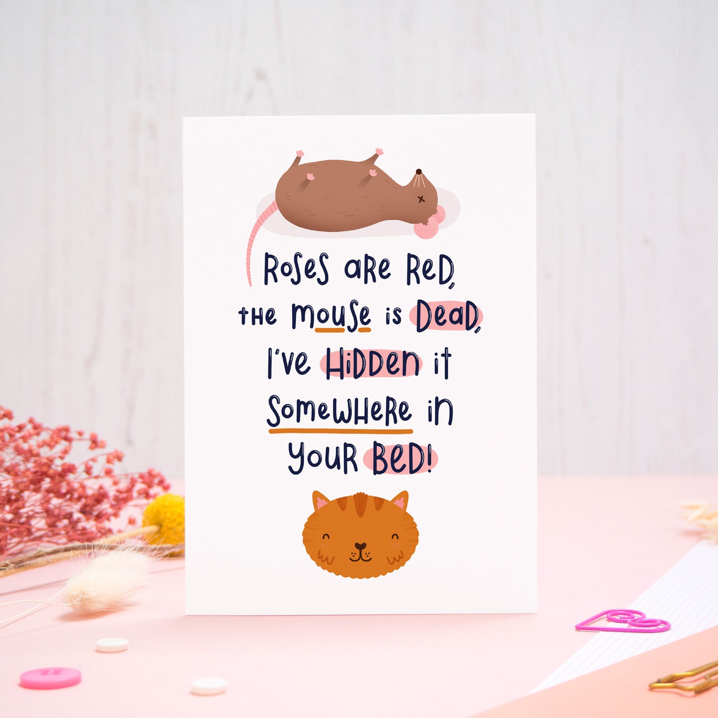 A Valentine’s ‘from the cat’ card featuring a ‘roses are red’ rhyme, photographed stood on a pink and white background with floral props, paper clips, and buttons. 