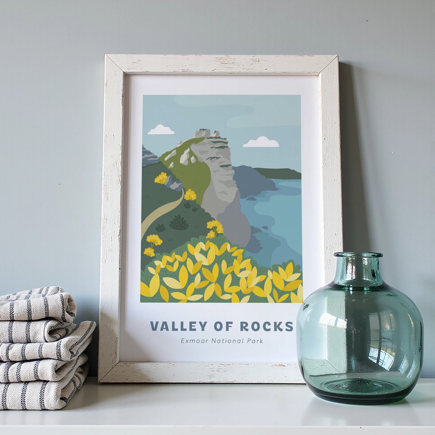 Valley of Rocks, Exmoor National Park art print in a white frame on a light grey background with a pile of cloths and blue vase.