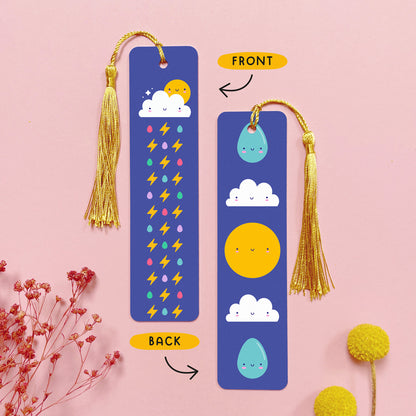 An image depicting the front and the back of the double sided metal bookmark which has been photographed on a pink background with dried flowers. Each bookmark has a gold tassel. On the front of the bookmark is a cloud, sun and colourful raindrops with lightning bolts which runs the length of the bookmark. On the reverse are two raindrops, two clouds and a sun, all with happy faces.