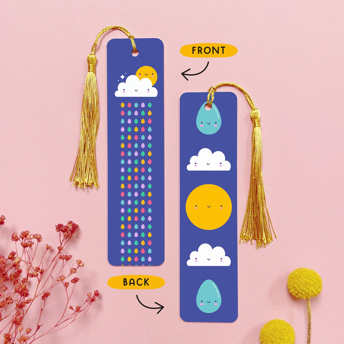 An image depicting the front and the back of the double sided metal bookmark which has been photographed on a pink background with dried flowers. Each bookmark has a gold tassel. On the front of the bookmark is a cloud, sun and a colourful rainbow raindrops which runs the length of the bookmark. On the reverse are two raindrops, two clouds and a sun, all with happy faces.