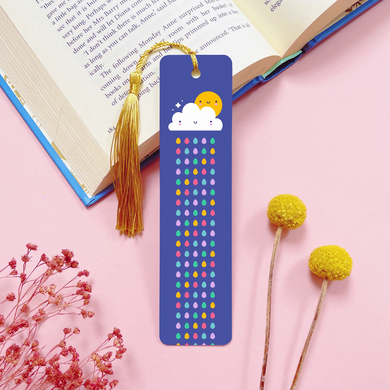 A raindrop themed, double sided, aluminium bookmark with a gold tassel which has been photographed on a pink background with an open book and dried flowers.