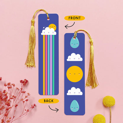 An image depicting the front and the back of the double sided metal bookmark which has been photographed on a pink background with dried flowers. Each bookmark has a gold tassel. On the front of the bookmark is a cloud, sun and a rainbow which runs the length of the bookmark. On the reverse are two raindrops, two clouds and a sun, all with happy faces.