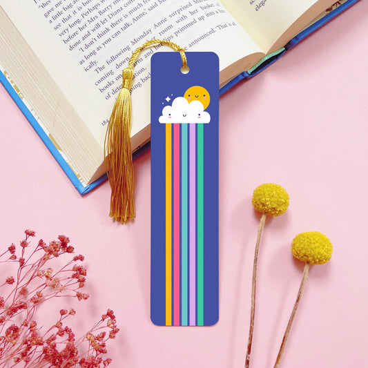 A rainbow themed, double sided, aluminium bookmark with a gold tassel which has been photographed on a pink background with an open book and dried flowers.