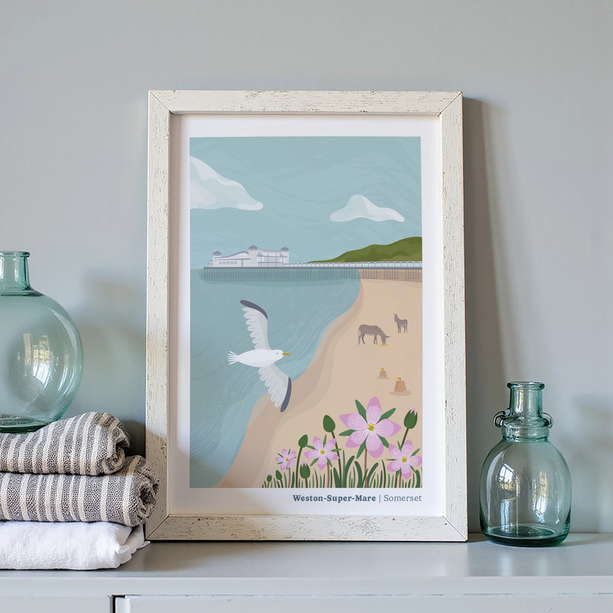 A framed art print featuring a coastal scene labeled 'Western-Super-Mare | Somerset' is displayed on a shelf. The print shows a seagull, donkeys, beach, flowers, and the sea, surrounded by folded towels and glass bottles.